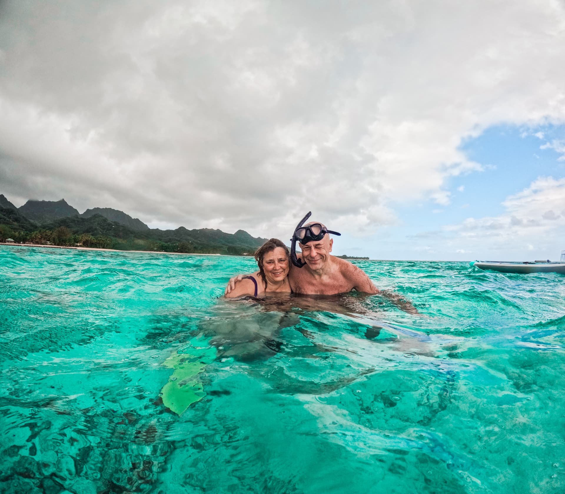 Two guests smiling in the turquoise Rarotonga lagoon with the mountains behind